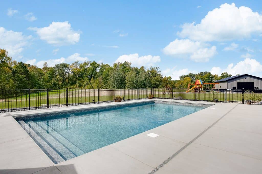A lush backyard with a modern concrete pool deck and safety fencing under a bright blue sky.
